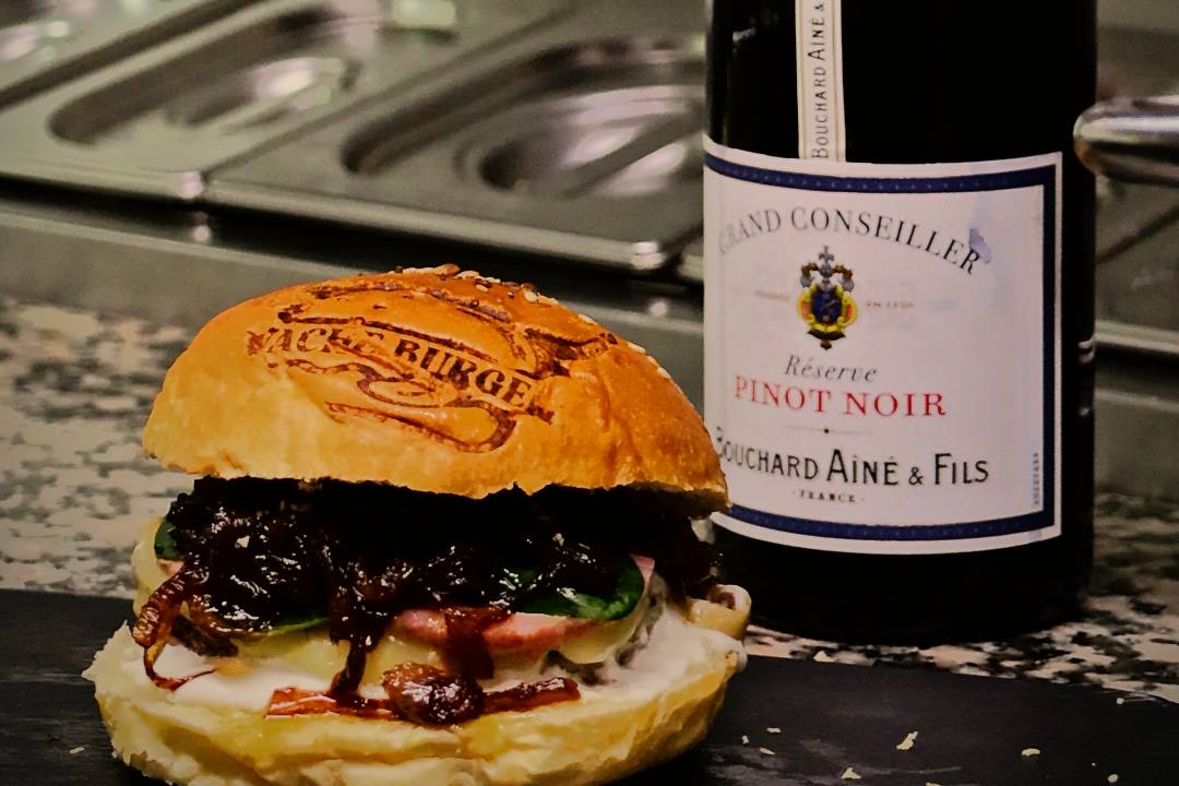 Un burger au confit d'oignon posé sur une ardoise, à côté d'une bouteille de pinot noir